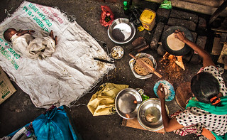 An overhead view of a woman squatting on a dirt floor, cooking over a brick oven. A baby wrapped in a dirty sheet sleeps on the floor nearby. 