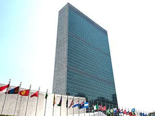 A tall glass and concrete structure fronted by the colorful flags of various nations.