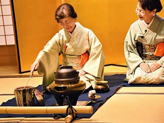 Two smiling women, wearing kimonos, kneel in front of a large teapot.