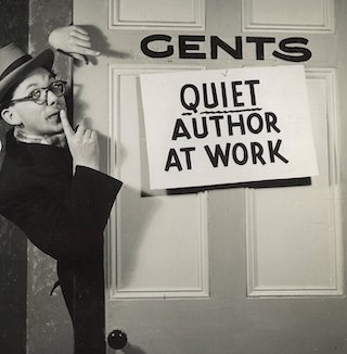Man with a finger to his lips peeking around a door with the sign "Quiet Author At Work" on it.