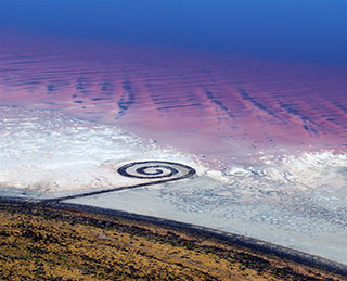 An aerial view of a spiral-shaped land art sculpture made out of mud, salt crystals, and balsalt rocks.