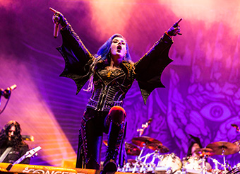 Female metal singer standing center stage holding a microphone in one hand with the drums and guitarist behind, both of her arms are raised revealing the studded batwing sleeves of her leather jacket.