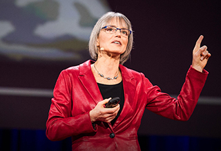 Photo of Nancy Kanwisher  standing on the dark TED stage in the middle of giving her talk, wearing glasses and a bright red blazer