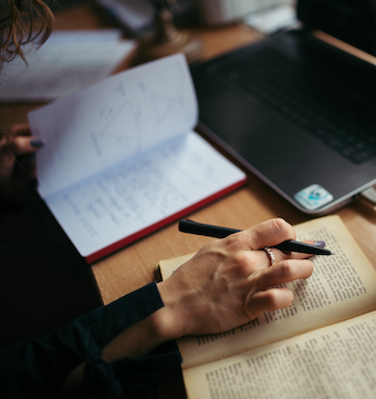 A woman sitting at a table with a laptop reading a book with her right hand with her left hand holding at a notebook.