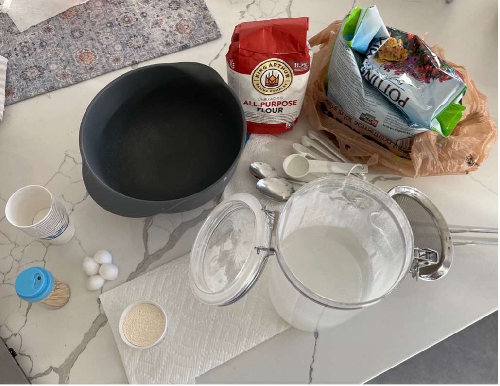 A table showing paper cups, toothpicks, cotton balls, flour, potting mix, a strainer, spoons, measuring spoons, a bowl, and two containers of powders.