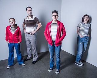 A man with a stern expression stands amongst three females: a preteen, a teenager, and an adult.  Each female wears jeans, sneakers, and a striped shirt.